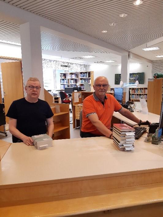 Kirjastonjohtaja Erkki Sirniö ja kirjastojärjestelijä Jouko Seppänen istuvat kirjaston lainaustiskissä. Menossa kuljetuksen tuomien varausten käsittely ja laittaminen varaushyllyyn.