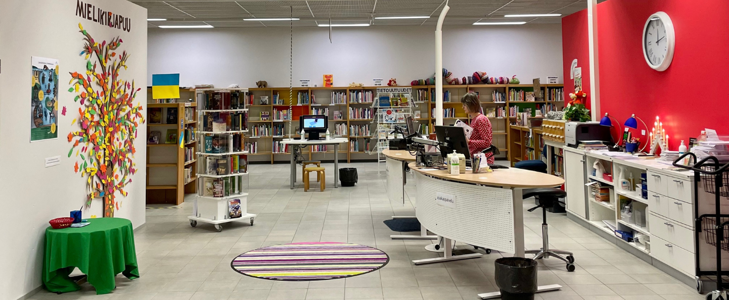 Kirjaston lastenosasto. Vasemmalla seinällä mielikirjapuu. Oikealla asiakaspalvelutiski, jonka takana työntekijä tietokoneella.