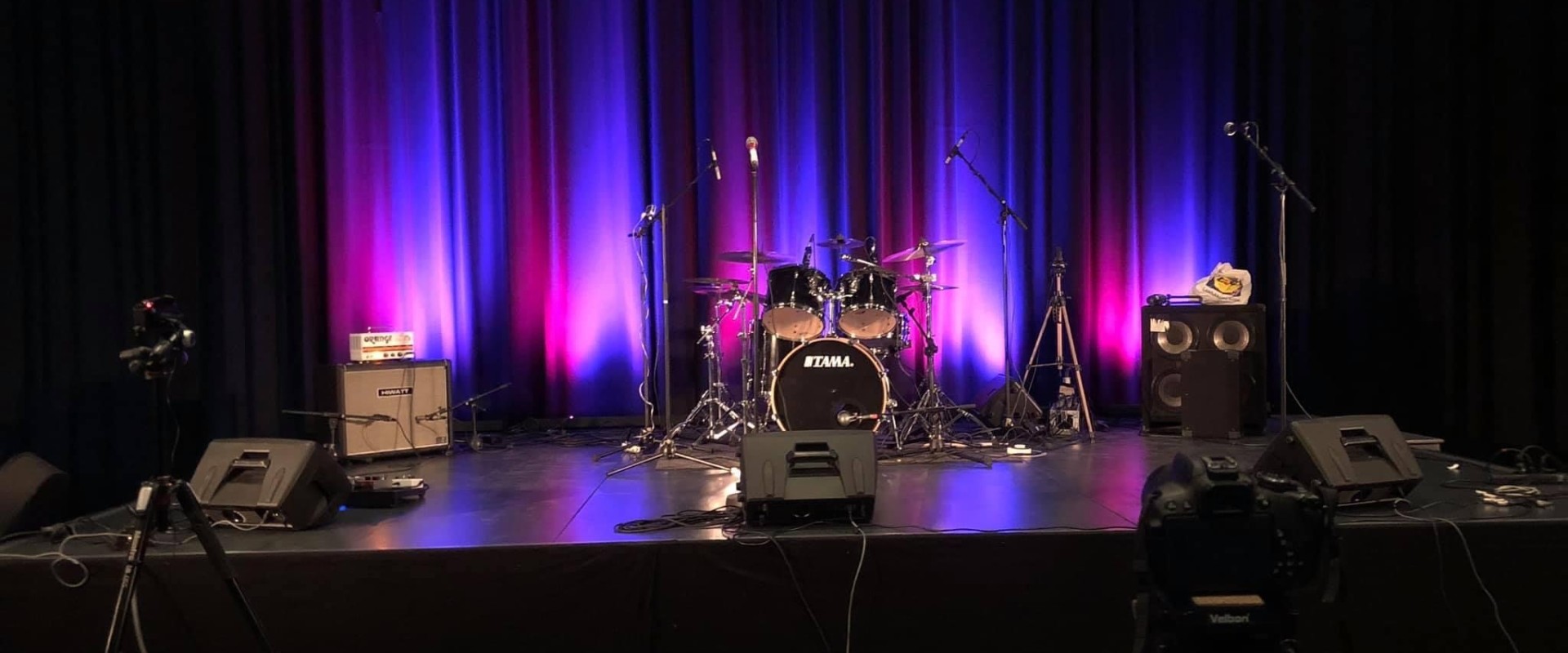 Tyhjä esiintymislava, johon aseteltu rummut, mikrofonit ja monitorit. Lava on valaistu dramaattisesti violetinsinisin sävyin.