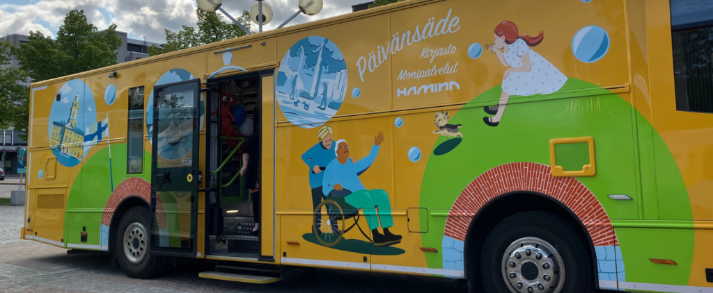 Haminan iloisen keltainen kirjasto auto, nimeltään päivänsäde. Kyljessä kuvitusta esimerkiksi Suomen lippu, pyörätuolissa oleva henkilö ja tyttö, joka puhaltaa saippuakuplia.