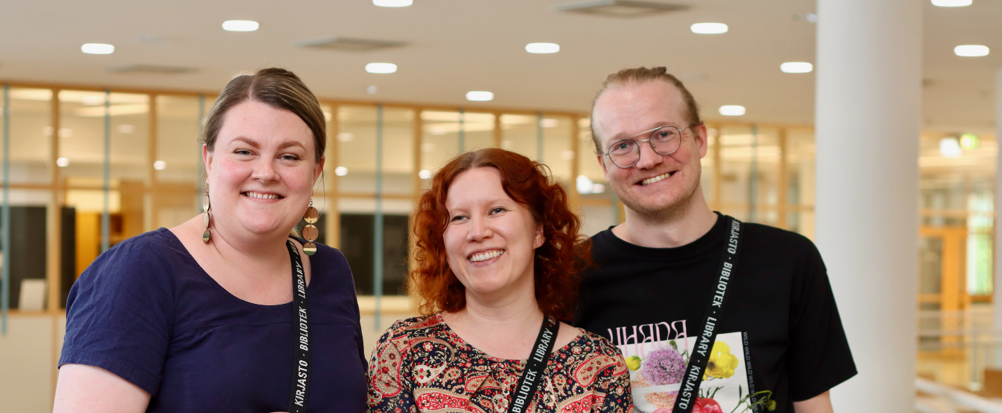 Koordinaattorit Noora, Hanna ja Pasi hymyilevät, kuva otettu valoisassa kirjastotilassa.