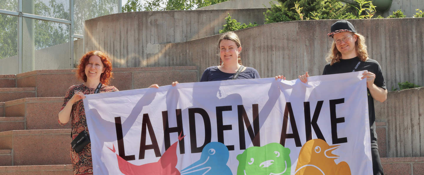 AKE-koordinaattorit Hanna, Noora ja Pasi kannattelevat AKE-logolla varustettua lakanaa Lahden kaupunginteatterin portailla.