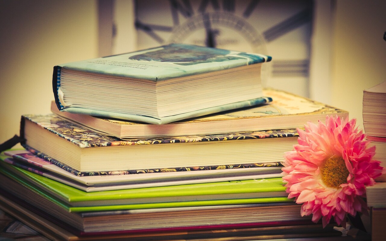 Pino kirjoja tasolla, kirjojen vieressä vaaleanpunainen kukka.