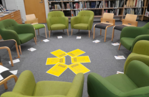 Vihreitä nojatuoleja aseteltu ympyrään harmaalle kokolattiamatolle kirjastotilassa. Keskellä tulosteina Erätauko-keskustelun pelisäännöt.