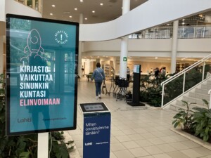 Vasemmalla infonäyttö, jolla kampanjaslogan Kirjasto vaikuttaa sinunkin kuntasi elinvoimaan. Infotaulun vieressä asiakaspalautteen keräämiseen tarkoitettu laite, jossa lukee Miten onnistuimme tänään. Taustalla Lahden pääkirjaston aulatila, jossa kävelee farkkutakkiin pukeutunut asiakas.